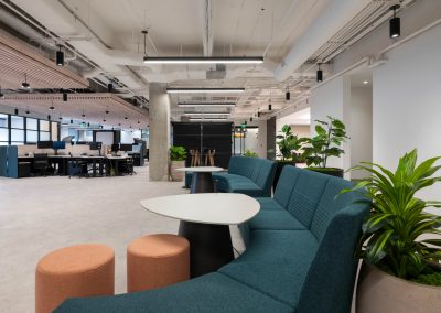Teal lounge seating, peach stools, and white tables surrounded by plants beside open office workstations.