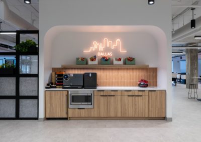 Office coffee station with wood cabinets, terracotta tile, snack shelves, and an illuminated Dallas skyline sign